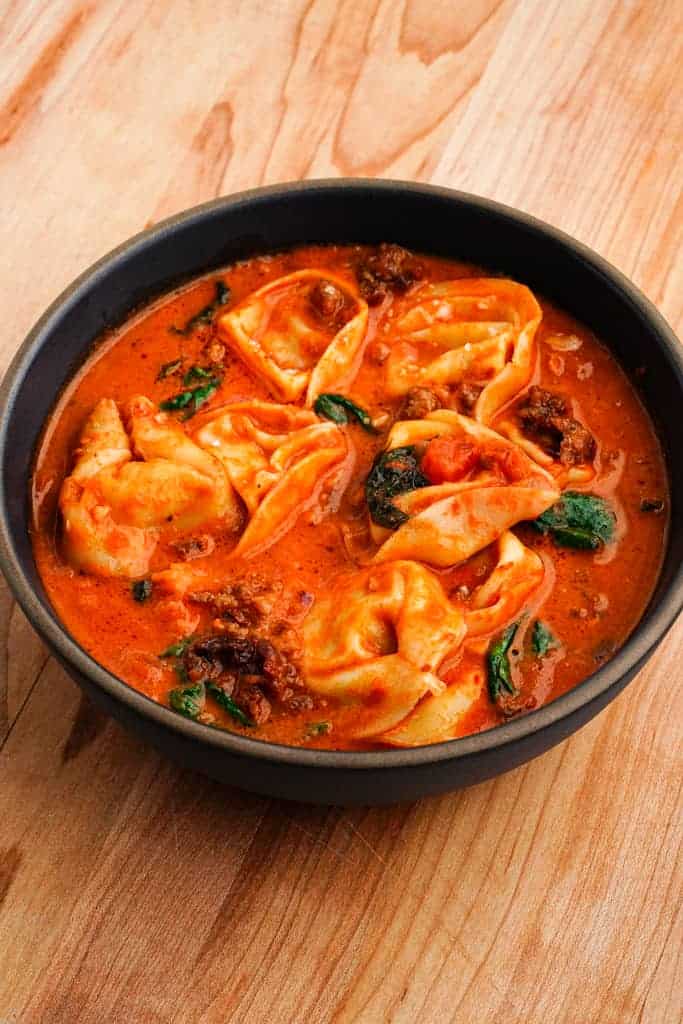 Bowl of creamy tortellini soup with sausage, spinach, and tomato broth, served on a wooden surface.