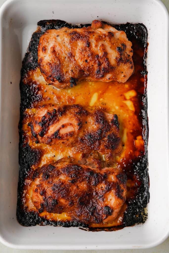 Roasted chicken thighs in a white baking dish surrounded by caramelized sauce and browned edges from oven roasting.